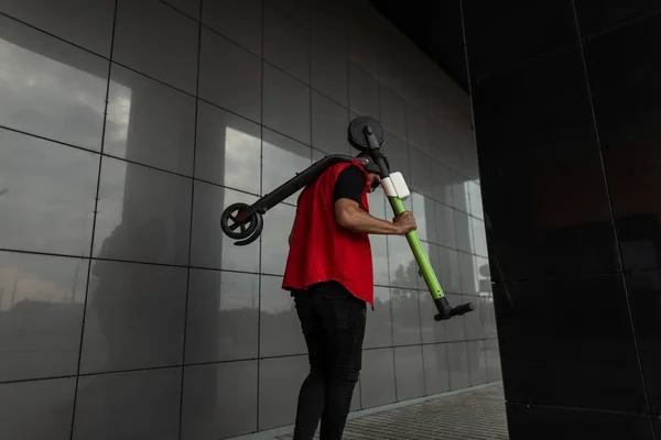 Vue arrière d'un Américain vêtu d'un gilet élégant rouge en jean noir tendance avec un scooter à l'épaule près d'un mur moderne noir dans la rue. Cool hipster guy promenades autour de la ville. Vue de l'arrière . — Photo