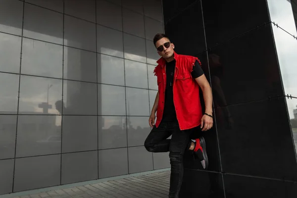 Der hippe junge Mann in roter Jeansweste und schwarzem T-Shirt in Jeans und Sonnenbrille posiert an einem Sommertag auf der Straße in der Nähe der Mauer. gutaussehendes urbanes Kerlmodell in der Stadt. Straßenmode. — Stockfoto