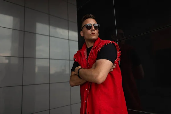 Ein junger attraktiver Hipster mit stylischer Sonnenbrille, T-Shirt und modischer roter Jeansweste posiert in der Nähe einer schwarzen Vintage-Wand in der Stadt. schöner moderner Kerl Modell. trendige Sommerbekleidung für Männer. — Stockfoto