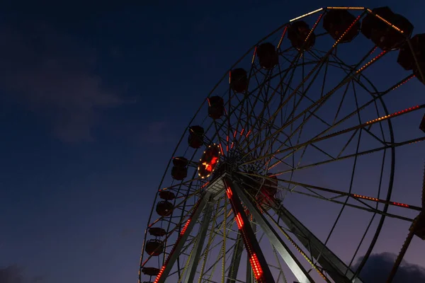 Part of a ferris wheel with lights of night illumination on a background a dark blue sky. Amusement park in Spain in the evening. View from below.