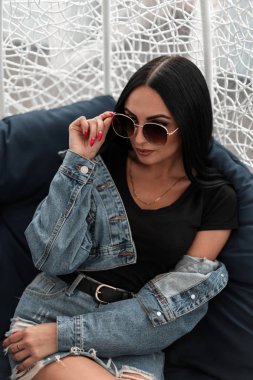 Çekici genç kadın modeli, bir yaz kafesinin terasında yumuşak bir sandalyede oturan şık güneş gözlüğünü düzeltiyor. Güzel hippi kotlu kız şehrin dışında dinleniyor..