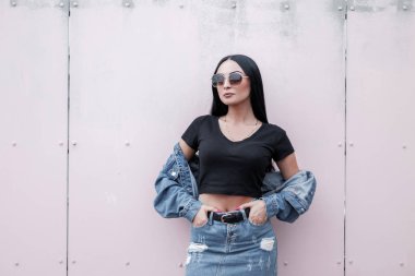Uzun güneş gözlüklü, esmer, güzel bir kadın. Klasik mavi etekli, şık, siyah bluzlu, kot ceketli. Şehirde modern bir binanın yanında duruyor. Moda mankeni..