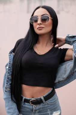 Güzel, esmer, uzun saçlı, modaya uygun kot ceketli, şık bir etekli, güneş gözlüklü, yaz günü açık havada bir binanın yanında poz veren güzel bir kadın. Amerikalı hippi kız.