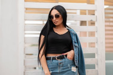 Şık siyah bluzlu, modaya uygun etekli, kot ceketli Amerikalı genç hippi kadın şehrin dışındaki beyaz ahşap bir duvarın yanında dinleniyor. Moda mankeni kız..