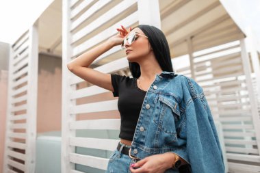 Modaya uygun, siyah uzun saçlı, siyah tişörtlü, şık kot elbiseli, şık güneş gözlüklü bir kadın manken. Güzel şehirli hippi kız klasik beyaz bir duvarın yanında poz veriyor..