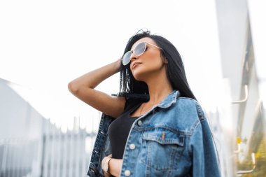 Modaya uygun güneş gözlüklü, şık bir kot ceketli, siyah tişörtlü hoş, genç hippi kadın saçları düzleştiriyor. Parlak bir sonbahar gününde şehirde dinlenen çekici bir esmer kız..