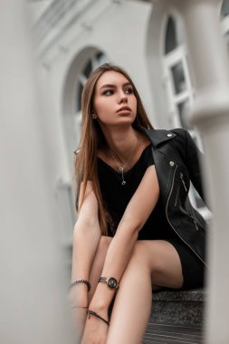 Şık siyah elbiseli zarif genç bir kadın beyaz bir binanın yanındaki basamaklarda oturuyor. Moda mankeni güzel bir kız. Moda kadın kıyafetleri. Sonbahar gündelik görünümü.