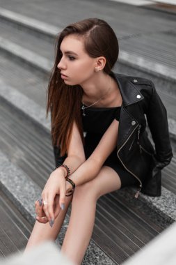 Modern beyaz bir binanın yanındaki klasik bir merdivende, modaya uygun siyah bir ceketle şık, kısa siyah elbiseli genç bir kadın model oturuyor. Çekici kız bir sonbahar günü şehirde dinlenir..