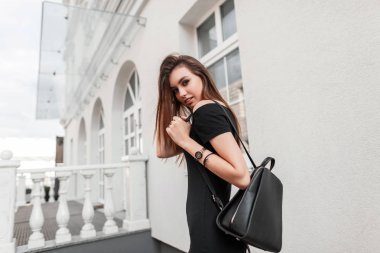 Zarif siyah bir elbise içinde, deri tarzı siyah bir sırt çantasıyla şehirde gezen oldukça güzel genç bir kadın manken. Şirin, şık bir kız sokakta yürümekten hoşlanıyor. Sokak gençliği tarzı.