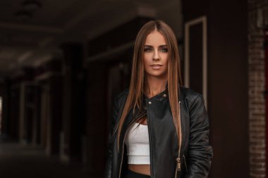 Çekici, doğal makyajlı, klasik deri ceketli, beyaz bluzlu, kahverengi saçlı, çekici bir genç kadın modeli karanlık bir odada duruyor..