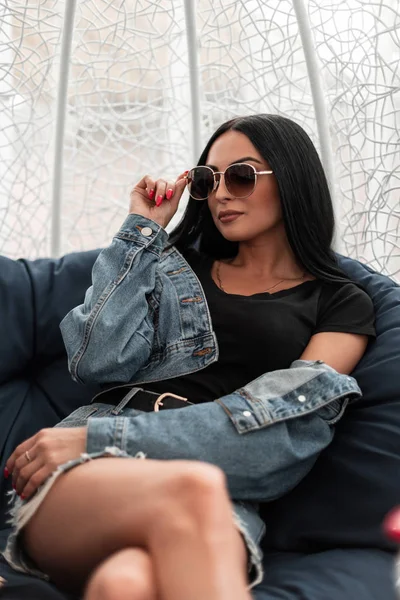 Amerikaanse jonge hipster vrouw in modieuze zonnebril in een zwart T-shirt in een stijlvolle denim jas in een rok ontspant zittend op een zachte fauteuil in een terras.Aantrekkelijk meisje rust buiten — Stockfoto
