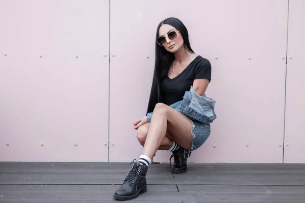 Mujer hipster bastante joven en gafas de sol en una ropa de mezclilla azul de moda en botas de cuero de otoño negro con estilo sentado cerca de una pared en la calle. Sexy chica urbana se relaja al aire libre. Moda callejera . — Foto de Stock