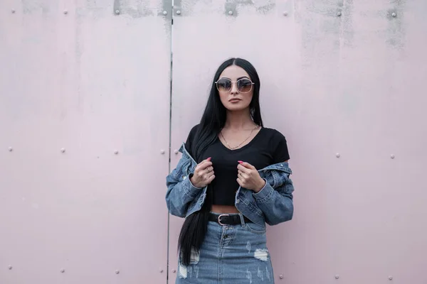 Moderne schöne junge Hipsterfrau mit stylischer schwarzer Sonnenbrille steht und richtet eine Jeansjacke in der Nähe einer Vintage-Wand. attraktive brünette Mädchen Modell in trendigen Kleidern posiert im Freien. — Stockfoto