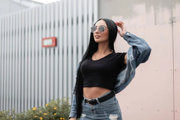 Model of a happy young woman with cute smile with black hair in a trendy black t-shirt in fashionable jeans jacket in a stylish sunglasses. Cheerful girl hipster rests near a vintage wall outdoors. — Stock Photo, Image