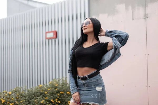 Joven morena con estilo alegre en una chaqueta de mezclilla de moda en una falda vintage en un top negro en gafas de sol posando cerca de una pared en la calle. Feliz chica hipster descansando al aire libre. Estilo juvenil . — Foto de Stock