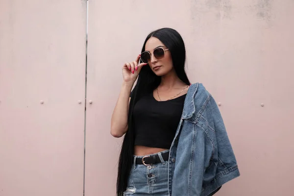 Atraktivní mladá brunetka s módní černou střechou ve stylové džínové bundě v retro modré džínové sukni pózující venku u budovy. Krásná hipster dívka narovnává sluneční brýle. — Stock fotografie