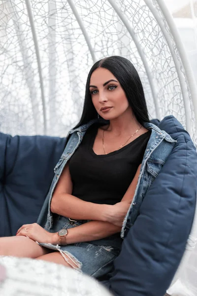 Ziemlich modisch schöne junge brünette Frau in einer stylischen Jeansjacke in einem T-Shirt sitzt auf einem Vintage-weichen Stuhl auf der Terrasse in einem Sommercafé. urbanes Hipster-Mädchen ruht in der Stadt. — Stockfoto