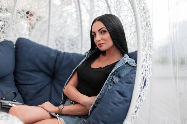 Trendy attraktive junge Frau Mode-Modell in modischen Jeanskleidung mit langen schwarzen Haaren entspannt auf einem Vintage-Sessel auf der Terrasse eines Sommercafés in der Stadt sitzen. schöne Mädchen im Freien — Stockfoto