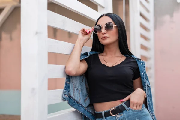 Urban stylish νεαρή γυναίκα hipster σε ένα μοντέρνο μαύρο top σε ένα μοντέρνο denim σακάκι σε μια φούστα σε σκούρα γυαλιά ηλίου ποζάρουν κοντά σε ένα λευκό ξύλινο τοίχο στην πόλη. Ελκυστικό κορίτσι μόδας μοντέλο σε εξωτερικούς χώρους. — Φωτογραφία Αρχείου