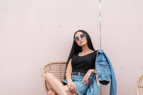 Urbain jeune femme brune à la mode dans des lunettes de soleil à la mode dans des vêtements en denim bleu élégant est assis sur une chaise de paille près d'un mur rose dans la ville. Attrayant hipster fille se détend à l'extérieur.Style de jeunesse — Photo