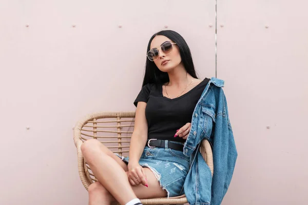 Όμορφη νεαρή γυναίκα με μαύρο μπλουζάκι σε τζιν σακάκι με γυαλιά ηλίου σε φούστα που ποζάρει σε ψάθινη καρέκλα κοντά στον ροζ τοίχο σε υπαίθριο καφέ. Μοντέρνο hipster κορίτσι.Μοντέρνο εποχιακά γυναικεία ρούχα — Φωτογραφία Αρχείου