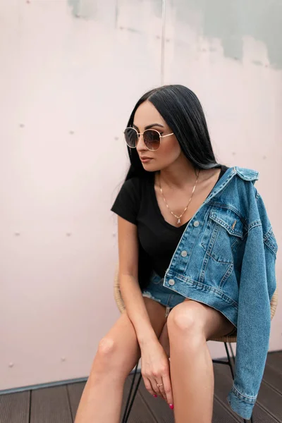 Gyönyörű fiatal barna nő divatos napszemüveg elegáns kék farmer ruhák ül egy széken, közel egy vintage rózsaszín fal a városban. Elég vonzó hipszter lány lazít a szabadban. Stílus. — Stock Fotó