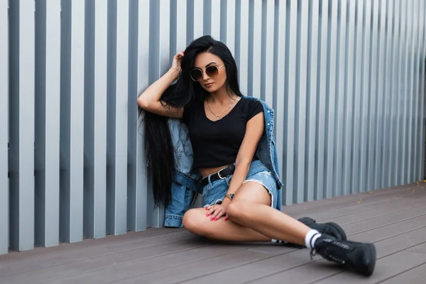 Eine junge Hipsterfrau mit Sonnenbrille sitzt in einer trendigen blauen Jeansjacke in stylischen schwarzen Lederstiefeln im Rock an einer silbernen Metallwand auf der Straße. Amerikanerin. Moderne Jugendmode. — Stockfoto