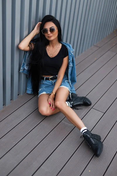 Trendy young woman hipster with long hair in fashionable jeans clothes in stylish black leather boots sits in the city near a silver metal wall. American girl rests outdoors. Stylish women's clothing. — Stock Photo, Image