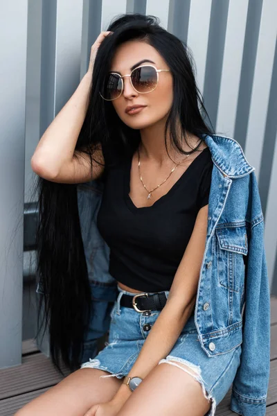 Elég vonzó fiatal barna nő egy elegáns fekete felső divatos farmer kabát napszemüvegben nyugszik közelében egy fém fal a szabadban. Trendy hipster lány modell élvezi a nyaralás a városban. — Stock Fotó
