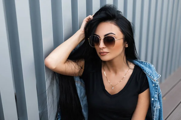 Όμορφη νεαρή μελαχρινή γυναίκα σε ένα μοντέρνο μαύρο t-shirt σε ένα μοντέρνο τζιν σακάκι με γυαλιά ηλίου στηρίζεται κοντά σε ένα μεταλλικό τοίχο σε εξωτερικούς χώρους. Σέξι hipster κορίτσι μοντέλο απολαμβάνει ένα Σαββατοκύριακο στην πόλη.Στυλ — Φωτογραφία Αρχείου