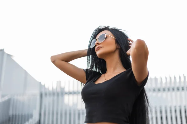 Belle jeune femme mannequin de mode avec des cheveux noirs chics dans un t-shirt noir à la mode dans des lunettes de soleil sombres élégantes posant en plein air dans la ville. Jolie fille hipster sexy dans la rue profite du week-end . — Photo