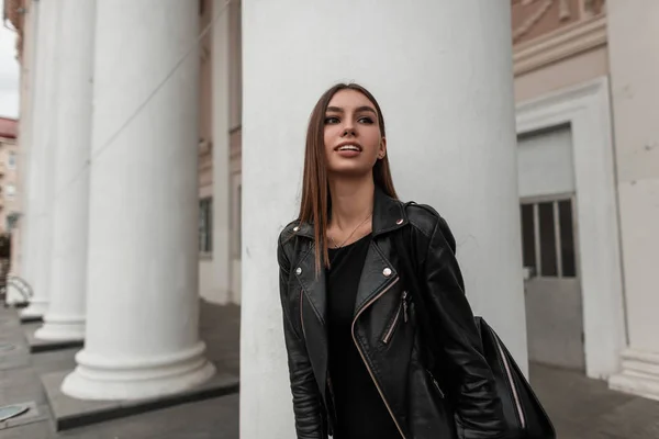 Jolie jeune femme urbaine dans une veste en cuir noir à la mode dans une robe élégante avec un sac élégant se tient près d'une colonne blanche vintage dans la rue. Jolie fille mignonne. Automne look décontracté . — Photo