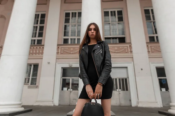 Urbain attrayant jeune femme aux cheveux bruns avec une veste élégante en cuir dans une robe noire avec un sac noir à la mode debout près d'un bâtiment vintage avec des colonnes. Fille promenades autour de la ville . — Photo
