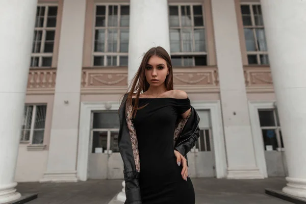 Jovem mulher sexy europeia em um vestido preto elegante em uma jaqueta de couro elegante com cabelo castanho posando na cidade perto de um edifício vintage. Urban muito atraente modelo de moda menina ao ar livre . — Fotografia de Stock