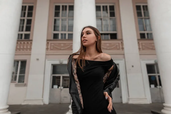 Elegante mooie jonge vrouw model in een modieuze jurk met blote schouders in een lederen zwarte stijlvolle jas poseren buiten in de buurt van een vintage gebouw. Sexy trendy aantrekkelijk meisje. Jongerenstijl. — Stockfoto