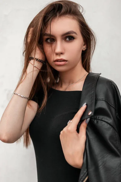 Pretty sensual young woman straightens hair. Cute attractive girl with beautiful eyes with sexy lips in a fashionable leather jacket in a stylish dress posing near a vintage white wall in the city. — Stock Photo, Image