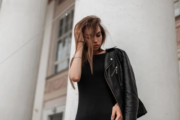 Mujer joven de moda bastante sexy endereza el cabello al aire libre. Modelo de chica atractiva en una chaqueta de cuero de moda en un elegante vestido negro posando en la ciudad cerca de un edificio vintage con columnas . —  Fotos de Stock