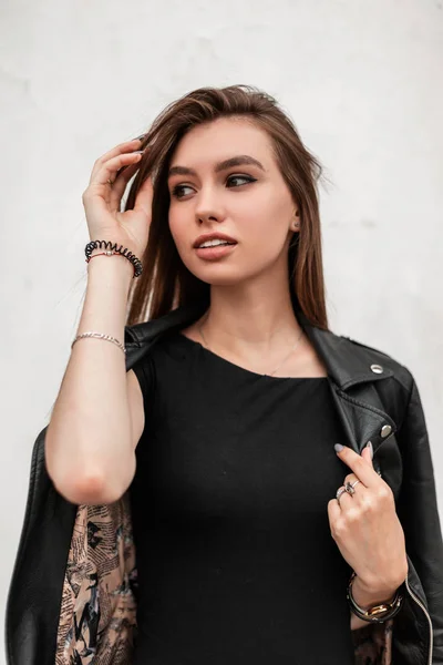Modern sensual young woman straightens hair. Trendy attractive girl with beautiful eyes with sexy lips in a fashionable leather jacket in a stylish dress posing near a vintage white wall in the city. — Stock Photo, Image