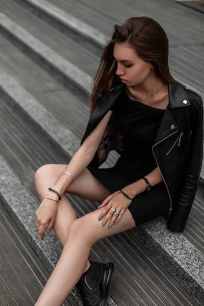 Mujer joven de moda en un vestido elegante en una chaqueta de cuero de moda en botas negras de otoño se relaja sentado en una escalera de piedra al aire libre. Moderna modelo de chica urbana descansando en la ciudad. Moda juvenil . —  Fotos de Stock