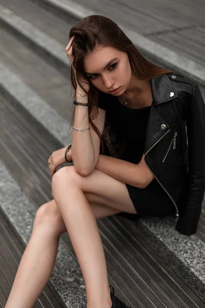 Sexy muito elegante jovem mulher no outono elegante roupas pretas em botas de couro vintage está descansando sentado em degraus de pedra na rua. Urbano bonito modelo de menina moderna relaxa ao ar livre . — Fotografia de Stock