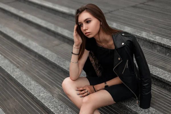 Jovem mulher atraente em roupas pretas elegantes com cabelo castanho posando sentado em uma escada de pedra na cidade. Bonito modelo de menina da moda descansando em um dia de outono ao ar livre. Roupas femininas na moda . — Fotografia de Stock