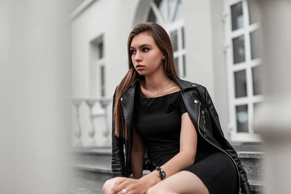 Modelo de mulher jovem europeu em um vestido preto curto elegante em uma jaqueta de couro preto elegante senta-se em uma escadaria vintage perto de um edifício branco moderno. Menina atraente descansa na cidade em um dia de outono — Fotografia de Stock