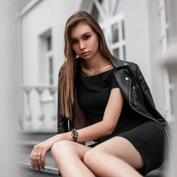 Atractiva joven europea con una chaqueta de cuero de moda en un elegante vestido negro se sienta en los escalones cerca de un edificio blanco vintage en la ciudad. Elegante modelo de chica moderna es relajante al aire libre . — Foto de Stock