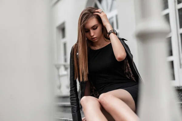Elegante modello giovane donna in un elegante abito corto nero in una giacca di pelle nera alla moda siede su una scala d'epoca vicino a un moderno edificio bianco. Ragazza moderna riposa in città in una giornata autunnale — Foto Stock