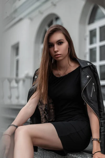 Cute beautiful young woman in a black short elegant dress in a stylish leather jacket sits on the stone steps near a vintage white building in the city. Fashionable urban girl relaxes on the street. — Stock Photo, Image