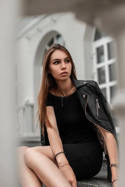 Modelo linda mujer joven de moda en un vestido corto y elegante en una chaqueta de cuero elegante se sienta en los escalones de piedra cerca de un edificio blanco vintage en un día de otoño. Chica urbana delgada de moda es descansa al aire libre — Foto de Stock