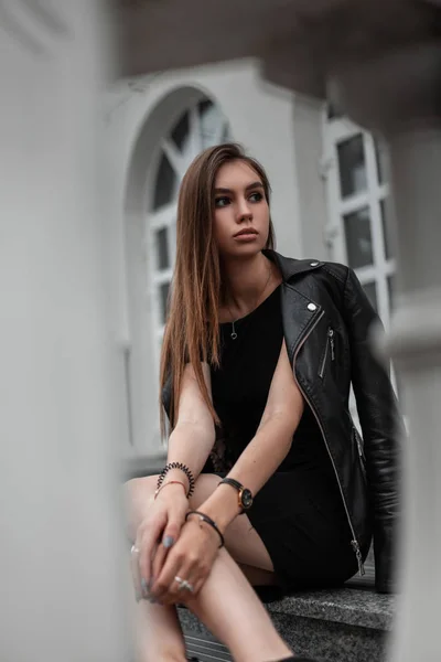 Młoda dość stylowa kobieta w czarnej modnej sukience w modnej skórzanej jesiennej kurtce relaksuje się na świeżym powietrzu w pobliżu zabytkowego budynku. Atrakcyjny model uroczej dziewczyny spoczywa na kamiennych schodach. Jesienny styl. — Zdjęcie stockowe