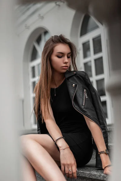 Belle jeune femme aux cheveux longs marron chic dans une robe élégante noire dans une veste vintage en cuir noir posant près d'un bâtiment moderne blanc. Mignon modèle de fille urbaine est assis sur les marches vintage . — Photo