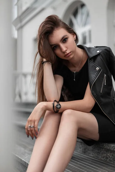 A jovem europeia atraente em uma jaqueta na moda de couro em um vestido preto senta-se nos degraus perto de um edifício branco vintage na cidade. Modelo de menina moderna elegante é relaxante ao ar livre. Moda . — Fotografia de Stock