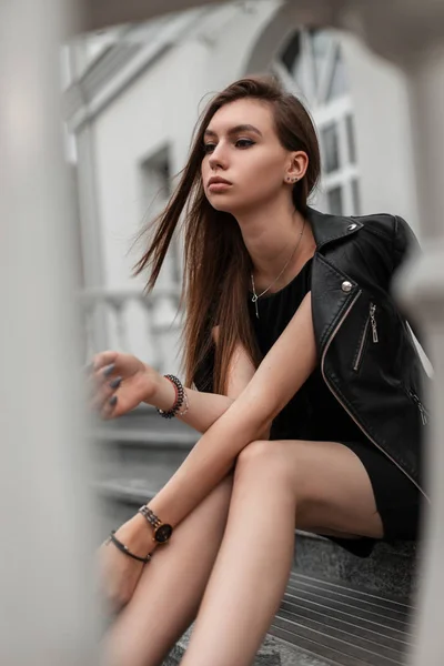 Modelo europeo de mujer joven en un elegante vestido negro corto en una elegante chaqueta de cuero negro se encuentra en una escalera de piedra cerca de un moderno edificio blanco. Hermosa chica descansa en la ciudad en un día de otoño . —  Fotos de Stock